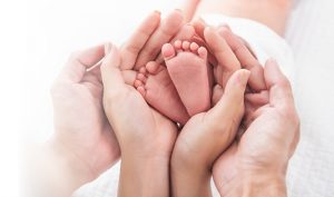 Eine Gruppe erwachsener Hände hält sanft die winzigen Füße eines Neugeborenen. Die Zehen des Babys sind gekrümmt und die Szene ist vor einem weichen, weißen Hintergrund angesiedelt, wodurch eine zarte und behütende Atmosphäre entsteht.