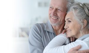 Ein älteres Paar lächelt und umarmt sich eng. Beide haben graues Haar; der Mann trägt einen grauen Pullover und die Frau ist weiß gekleidet. Der Hintergrund ist sanft verschwommen und strahlt eine helle, heitere Atmosphäre aus.