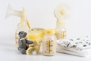 Eine Sammlung von Babyartikeln, darunter eine Milchpumpe, drei mit Milch gefüllte Flaschen, ein gestreifter Teddybär, ein Schnuller und ein Stück weißes Tuch mit Pfotenabdrücken, angeordnet auf einem weißen Hintergrund.