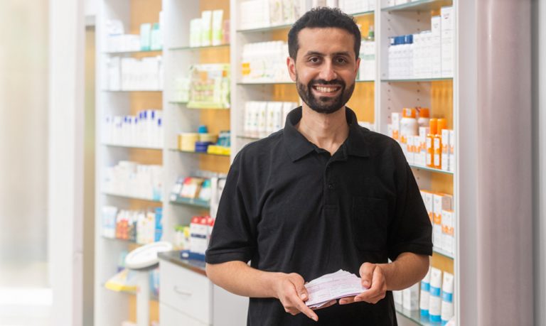 Ein Mann in einem schwarzen Poloshirt lächelt, während er in einer Apotheke eine Packung Medikamente hält. Hinter ihm sind Regale mit verschiedenen pharmazeutischen Produkten und Gesundheitsartikeln gefüllt. Die Umgebung ist hell und ordentlich.