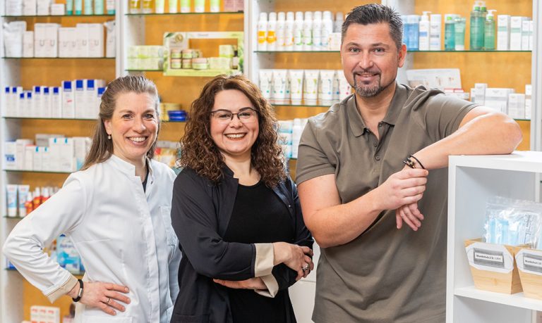 Drei lächelnde Menschen stehen in einer Apotheke. Links steht eine Frau im weißen Kittel, in der Mitte eine weitere Frau mit lockigem Haar und Brille und rechts steht ein Mann mit Bart, der an einem Regal lehnt. Im Hintergrund sind Regale mit verschiedenen Produkten zu sehen.