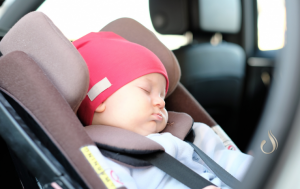 Ein Baby mit roter Mütze und weißem Hemd schläft friedlich in einem Autositz in einem Fahrzeug. Die fest befestigten Gurte gewährleisten die Sicherheit des Babys und entsprechen den Grundsätzen der Reisemedizin. Der schwarze Innenraum des Autos unterstreicht, wie zufrieden und bequem das Baby wirkt.
