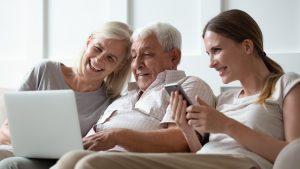 Ein lächelnder älterer Mann sitzt zwischen zwei Frauen und schaut auf einen Laptop-Bildschirm. Die Frau links hat graues Haar und die Frau rechts, die ein Smartphone hält, hat braunes Haar. Sie scheinen einen Moment zu genießen, in dem sie etwas über Apotheken – Neu-Isenburg & Bischofsheim austauschen oder lehren, und sitzen gemütlich auf einem Sofa.