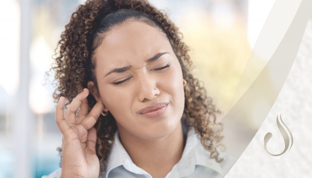 Frau fasst sich mit Finger ans Ohr. Ihr Gesicht ist verzogen. Zum Thema Was bedeutet Rauschen im Ohr
