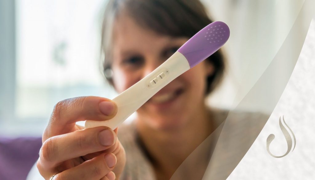 Glückliche Frau zeigt einen positiven Schwangerschaftstest in die Kamera zum Thema Nahrungsergänzungsmittel bei Kinderwunsch