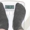 Füße in dunklen Socken auf einer Waage, die in der Anzeige Help anzeigt zum Thema Adipositas - Was ist das?